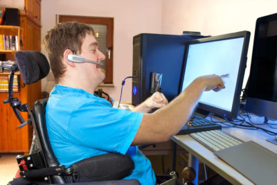 Man in wheelchair and talking on headset points at computer screen.